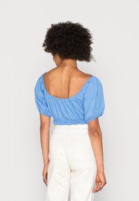 Blusa azul corta com mangas bufantes e um decote largo. A parte de trás apresenta um design em scoop baixo, combinada com calças de cintura alta de cor clara.