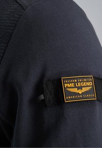 Écusson en tissu noir avec texte jaune « FREEDOM UNLIMITED PME LEGEND AMERICAN CLASSIC » attaché à un tissu texturé bleu marine foncé.