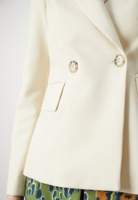 Krémový dvouřadý blazer s dvěma lesklými stříbrnými knoflíky, strukturovanými rameny a ostrými klopami, zkombinovaný s vzorovanými zelenými kalhotami.