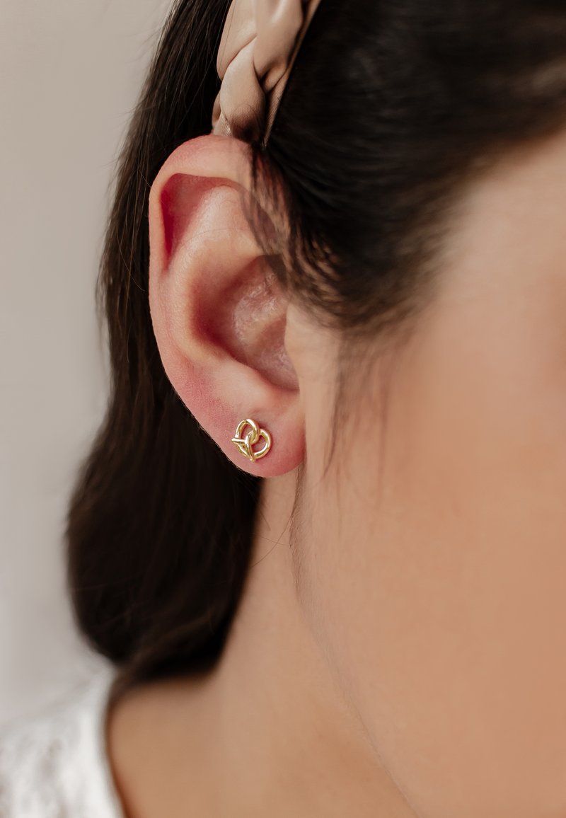 Pendiente de aro en forma de nudo dorado, de diseño pequeño y sencillo, con superficies lisas y un acabado pulido, llevado en un lóbulo de la oreja.