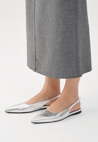 Zapatos planos tipo slingback en plata metálica, con puntera afilada, textura lisa, con cierre de hebilla en el tacón, combinados con una falda gris.