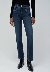 Jeans de denim azul oscuro con un diseño de pierna recta, que cuentan con dos bolsillos frontales, acentos de botón dorado y costuras en contraste. Llevados con botas negras.