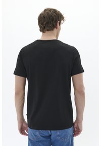 Melna kokvilnas T-krekls ar īsām piedurknēm un apaļu kakla izgriezumu, skatīts no aizmugures. Tam ir vienkāršs dizains un taisna apakšmala.