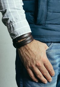 Brown leather bracelet with braided detail, worn on a hand, featuring a textured surface. Casual denim and a blue puffer vest in the background.