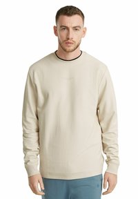 Beige sweatshirt with a textured fabric, crew neckline, and long sleeves. Features subtle brand text in the center. Worn with light blue pants.