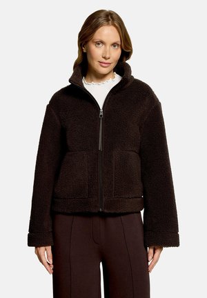 Femme portant une veste polaire zippée marron foncé avec col montant et poches avant, par-dessus un haut blanc à col à volants, debout devant un fond uni.