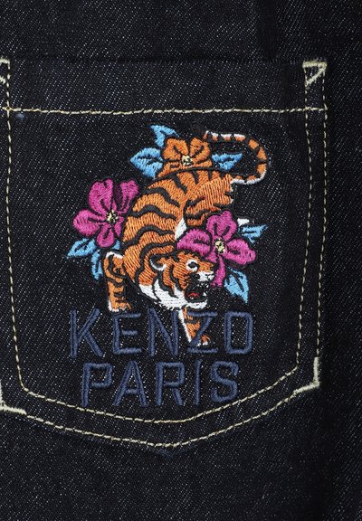 Un tissu en denim noir présente une patch avec un tigre brodé en orange et blanc, entouré de fleurs roses et bleues, et le texte "KENZO PARIS."