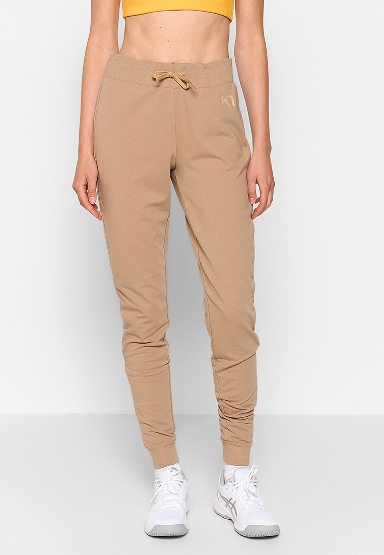 Slim-fit beiga joggers med elastisk midja och dragsko, bärs med vita sneakers och en gul croppad topp på en stående person.