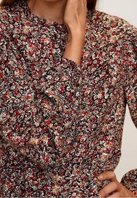 Bloes met bloemenprint, gekenmerkt door een zwarte achtergrond met rode, witte en gele bloemen. Lange mouwen met knoopdetails bij de kraag.