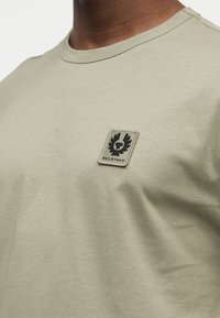 Κοντινό πλάνο ενός ατόμου που φοράει μια ανοιχτοπράσινη ελιάς κοντομάνικη μπλούζα με ένα μικρό μαύρο ετικέτα λογότυπου Belstaff στο αριστερό στήθος.