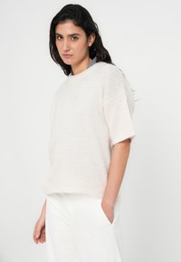 Ανοιχτό μπεζ πλεκτό πουλόβερ με κοντά μανίκια και άνετη εφαρμογή, από μαλακό, υφασμένο ύφασμα, φορεμένο πάνω από ανοιχτόχρωμο παντελόνι.