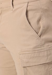 Shorts cargo beiges avec poches latérales, fabriqués dans un tissu doux et résistant. Présentent des coutures visibles et une poche cargo sécurisée sur le côté.