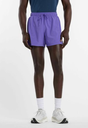 Uomo indossa pantaloncini sportivi viola, calzini bianchi, scarpe da corsa bianche e una maglietta blu, in piedi contro uno sfondo bianco semplice.