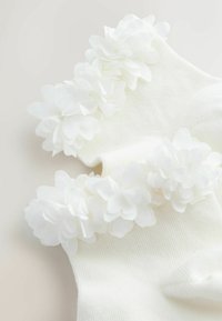 Fehér zokni bordázott mintával, a mandzsettán fehér szövetvirágokkal díszítve, puha és díszes részletet alkotva.