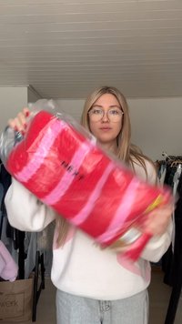 Coperta a righe rosse e rosa in confezione trasparente, con il nome del marchio "NEXT" stampato in nero. Texture morbida, forma rettangolare.