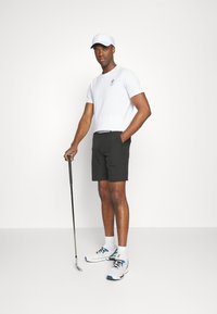 Hvit t-skjorte med grønt logo, svarte shorts, hvit caps og grå joggesko. Holder en golfklubbe, står foran en nøytral bakgrunn.