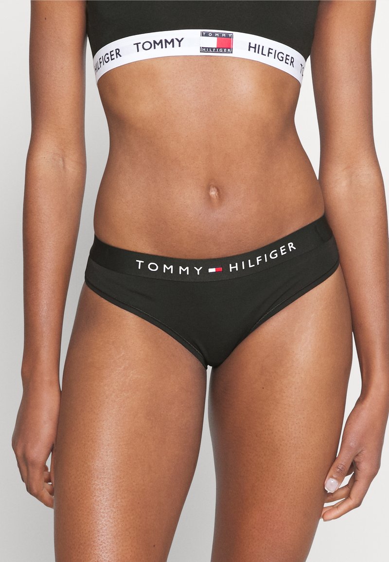 Culotte de bikini noire avec une large taille élastique arborant la marque "TOMMY HILFIGER" en blanc. Tissu lisse, coutures minimales et coupe ajustée.