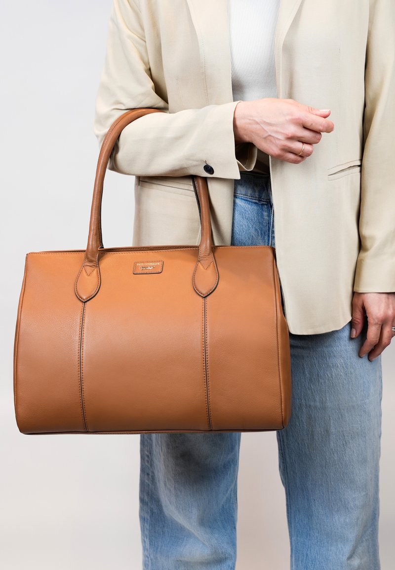 Sac fourre-tout en cuir tan avec une texture lisse, présentant deux poignées robustes et un logo en relief subtil. Forme structurée et lignes épurées.