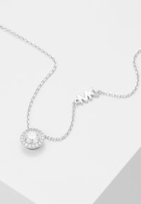 Collana in argento con un pendente circolare che presenta una pietra centrale trasparente circondata da cristalli più piccoli, contrassegnata con "MK."