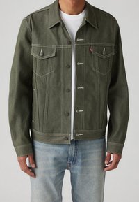 Olivgrön denimjacka med knappfram, bröstfickor, vit sömnad och kortad passform, i kombination med ljusblå jeans.