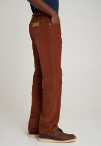 Rudos spalvos manžetinės kelnės su tiesia forma, turinčios austą diržo kilpą, priekinės kišenės ir galinė landynė. Derinamos su rudais batais.