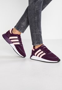 Lila sportskor med rosa randiga detaljer, överdel av tyg, snörning, och en vit mellansula på en strukturerad svart yttersula.