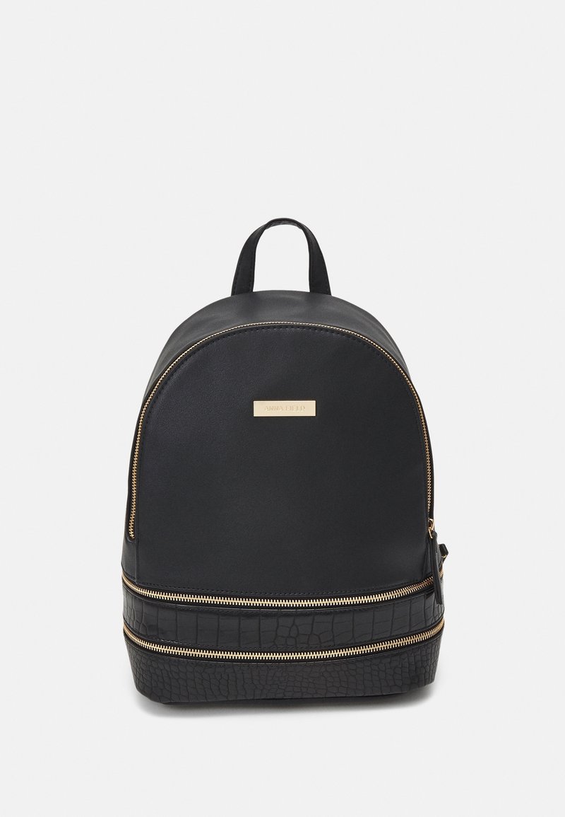 Sac à dos noir avec fermetures éclair dorées, une poignée supérieure et une section inférieure texturée, debout sur un fond gris clair uni.