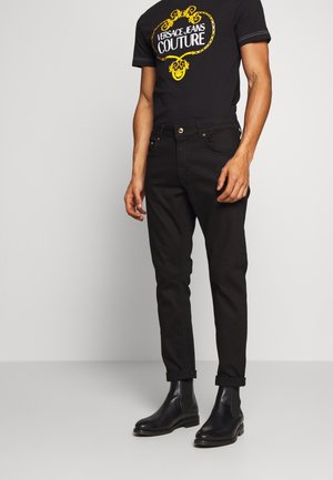 Homme portant un t-shirt noir Versace Jeans Couture, un pantalon noir retroussé et des bottines en cuir noir, debout devant un fond uni.