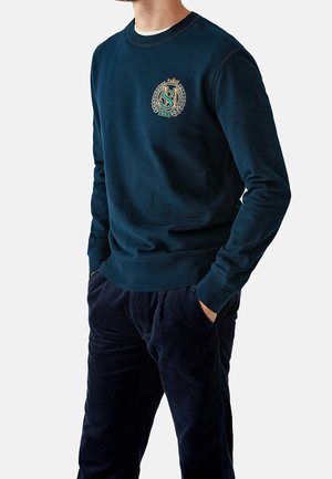 Marineblaues Sweatshirt mit Rundhalsausschnitt, versehen mit einem bestickten Logo und Lorbeerdesign. Weiche Textur, kombiniert mit marineblauen Cordhosen.