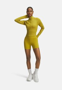 Vrouw staat in een mosterdgele lange mouwen zip-up top en bijpassende shorts met witte sneakers en sokken op een witte achtergrond.