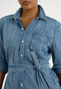 Detailný záber na osobu nosiacu svetlomodré rifľové šaty s gombíkmi vpredu, náprsným vreckom, vyhrnutými rukávmi a opaskom z rovnakého materiálu previazaným v páse.