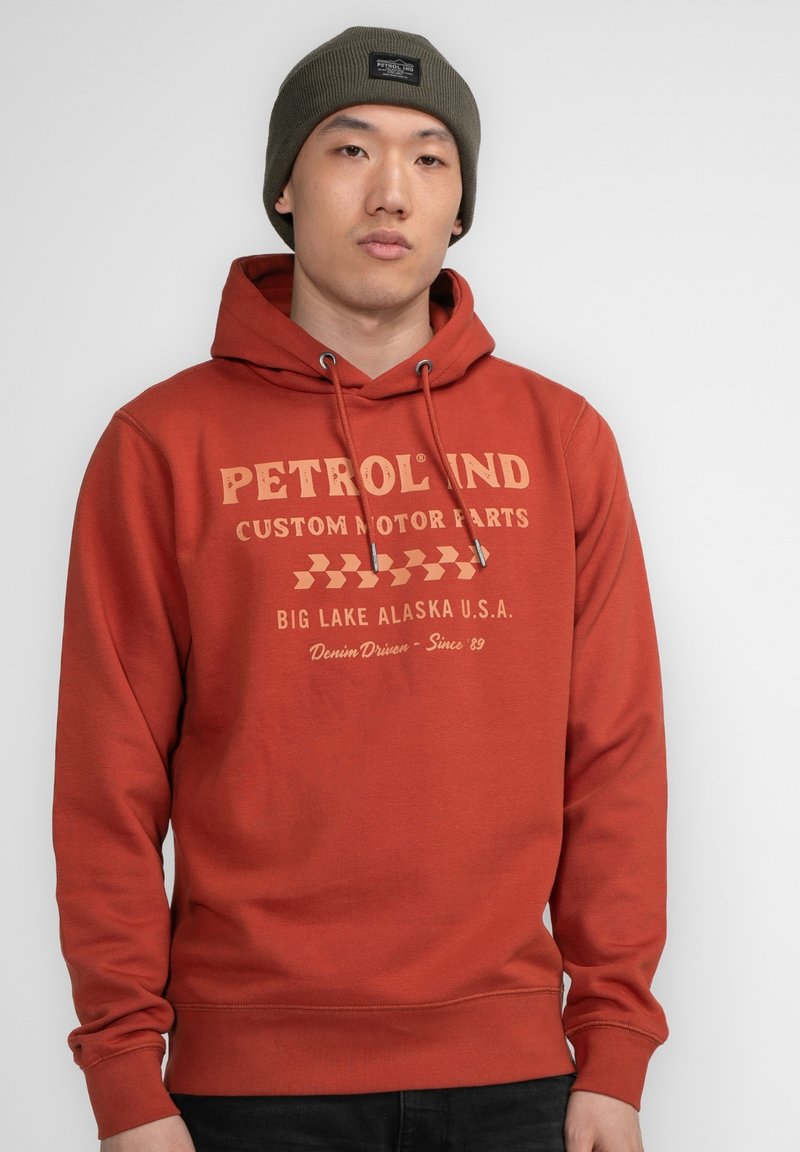 Felpa color ruggine con stampa grafica in crema. Presenta un cappuccio con cordino e polsini a coste. Indossata con un berretto oliva.