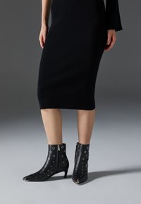 Botas negras de cuero hasta el tobillo con punta afilada, que presentan recortes circulares y cremalleras laterales. El tacón es moderado y texturizado.