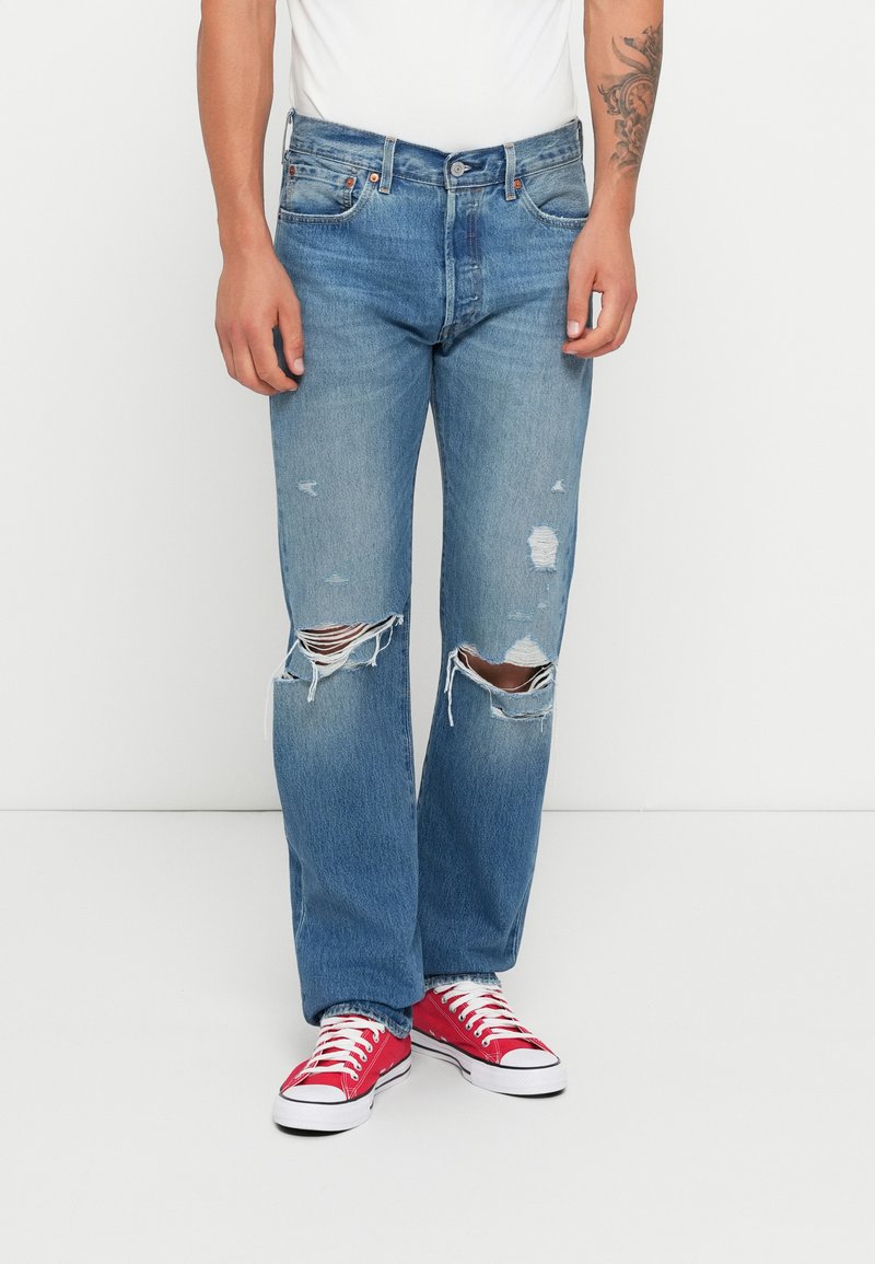 Homem a usar jeans azuis desgastados com joelhos rasgados, sapatilhas de lona vermelhas e uma camisa branca diante de um fundo branco simples.