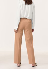 Pantalon beige à jambes larges et taille haute, doté de poches arrière et d'une texture de tissu lisse. Associé à un chemisier blanc à manches longues.