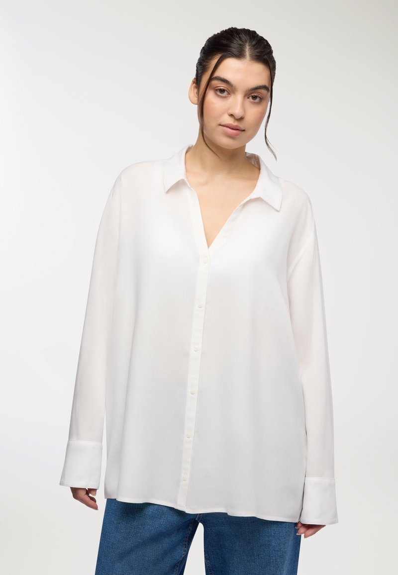 Camicia bianca con bottoni realizzata in tessuto leggero, dotata di scollatura a V, maniche lunghe e una vestibilità rilassata, abbinata a jeans in denim blu.