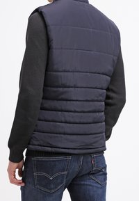 Granatowa pikowana kamizelka z wysokim kołnierzem, z pikowaną teksturą i czarnymi dzianinowymi rękawami. Noszona na ciemnoniebieskich jeansach.