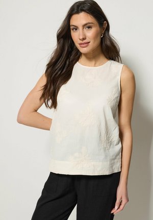 Donna con capelli lunghi e scuri che indossa una canotta crema con ricamo floreale e pantaloni neri, posando su uno sfondo chiaro.