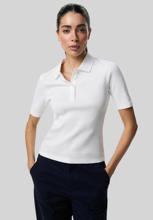 Kobieta o ciemnych włosach zaczesanych do tyłu, ubrana w biały polo z krótkim rękawem i ciemne spodnie, ręce w kieszeniach, neutralne tło.