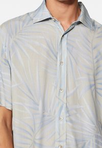 Camisa de manga corta en beige claro con estampado de hojas de palma azules, con cuello abotonado y botones claros en la parte delantera.