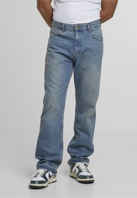 Spodná časť tela osoby oblečenej v svetlomodrých džínsoch s ošúchanými lemami a čierno-bielych teniskách, stojacej na jednoduchom svetlom pozadí.