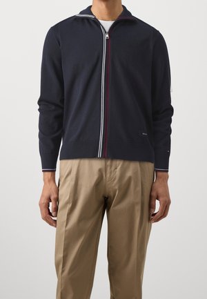 Man wearing a navy zip-up sweater with white and red trim, beige trousers, standing against a plain light background.