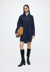 Rifľové košeľové šaty s dlhými rukávmi, náprsným vreckom, v tmavomodrej farbe. Kombinované s čiernymi koženými čižmami po kolená a hnedou taškou.