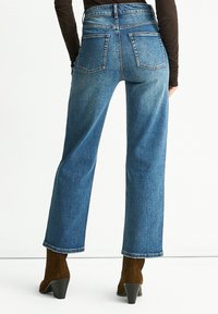 High-waisted blue denim jeans with a straight leg, featuring two back pockets and a raw edge hem, paired with brown heeled ankle boots.