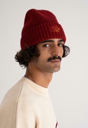 Czerwony prążkowany beanie z podwiniętym mankietem i złotym haftowanym logo. Teksturowany materiał dzianinowy kontrastuje z kremowym swetrem pod spodem.
