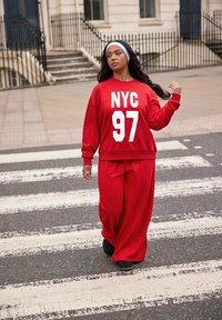 Sudadera roja de gran tamaño con "NYC 97" en blanco negrita, combinada con pantalones rojos holgados. Unos zapatos negros completan el conjunto deportivo en un paso de peatones.