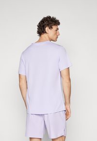 Šviesiai violetinė trumpomis rankovėmis marškinėliai su apykakle, dėvimi su atitinkamais violetiniais šortais. Audinys atrodo lygus ir lengvas.