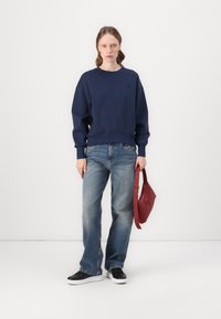 Mørk marineblå sweatshirt, avslappet passform med ribbestrikkede mansjetter, kombinert med lyseblå rett-snittjeans og svarte joggesko, mens hun holder en rød håndveske.