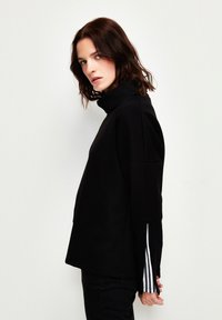 Maglione nero a collo alto con una vestibilità rilassata, fessure laterali e righe bianche a contrasto sui polsini. Texture morbida, design minimalista.