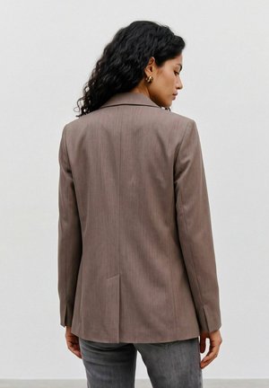 Mujer con cabello negro ondulado que lleva pendientes de aro dorados y una chaqueta marrón, de pie de espaldas sobre un fondo liso y claro.